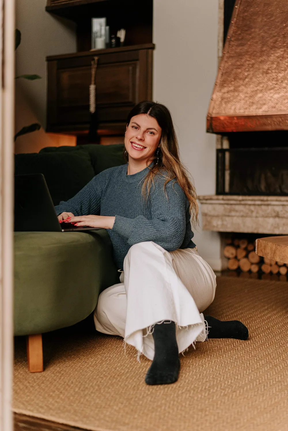 Adriana, fundadora da ArteNova, sentada no chão da sala, sorrindo e trabalhando no notebook